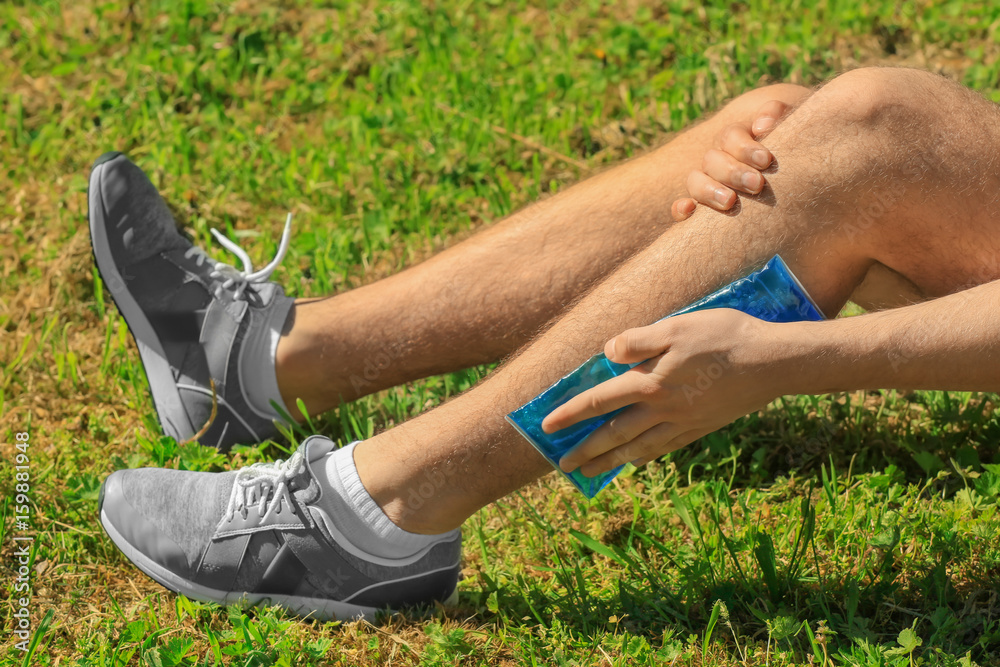 Young man applying cold compress to leg while sitting on grass outdoors, closeup Young man applying cold compress to leg while sitting on grass outdoors, closeup