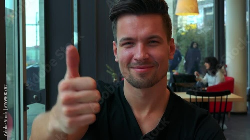 A young handsome man sits at table in a cafe and shows a thumb up to the camera with a smile - closeup photo