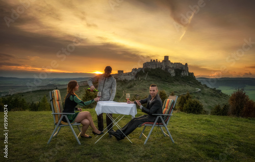 Party of three people drink red wine in nature under the ruins of a castle at sunset.