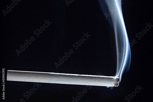 A completely white cigarette begins to smolder and smokes against a dark background. photo