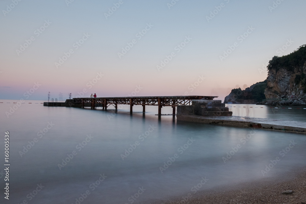 Fototapeta premium Wooden dock at the beach with blurred water at sunset and people silhouettes 