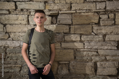 handsome man in glasses standing against a stone wall wearing a black t-shorts looking straight in to camera.