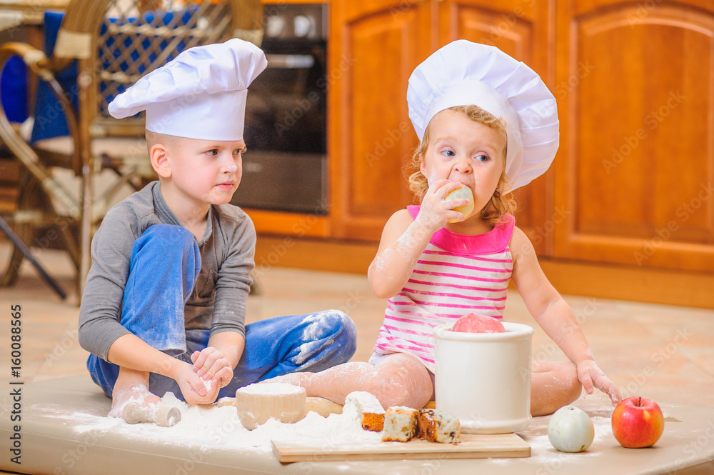 Two siblings - boy and girl - in chef's hats sitting on the kitchen floor soiled with flour, playing with food, making mess and having fun Two siblings - boy and girl - in chef's hats sitting on the kitchen floor soiled with flour, playing with food, making mess and having fun
