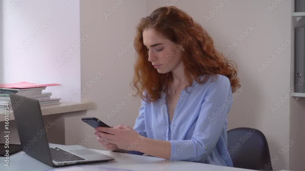 redheaded young woman messaging on smartphone. Redhead girl sitting at working place at home office. Smiling businesswoman using app on mobile phone. redheaded young woman messaging on smartphone. Redhead girl sitting at working place at home office. Smiling businesswoman using app on mobile phone.