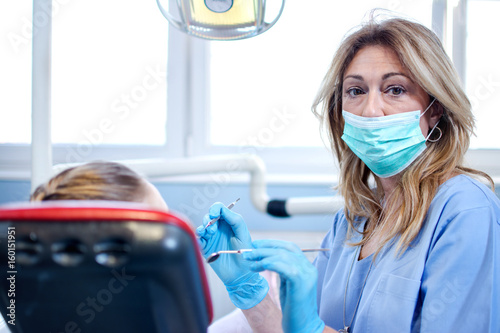 Portrait of middle age female dentist with protective mask holding dental tools and sitting near patient in dental office.