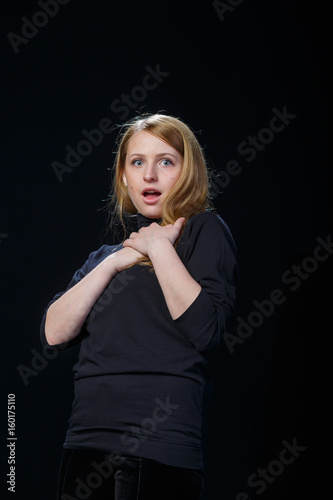 Surprised young blonde raised her hands to her chest against a black background