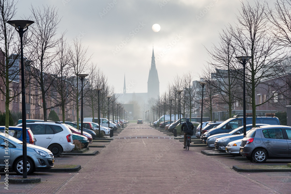 Provincial Holland. Cityscape With Solar Eclipse, Street Of Small Flanders Town With Gothic Church, Two Rows Of Cars And A Bicyclist. Solar Eclipse Under The Town