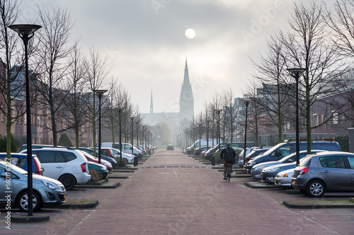 Obraz na płótnie Provincial Holland. Cityscape With Solar Eclipse, Street Of Small Flanders Town With Gothic Church, Two Rows Of Cars And A Bicyclist. Solar Eclipse Under The Town w salonie