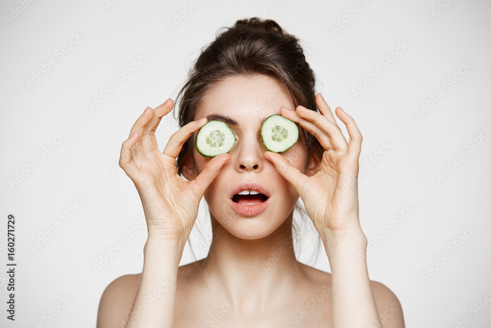Young beautiful nude girl with opened mouth hiding eyes behind cucumber slices over white background. Beauty skincare and cosmetology. Young beautiful nude girl with opened mouth hiding eyes behind cucumber slices over white background. Beauty skincare and cosmetology.