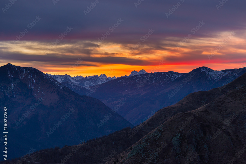 Colorful sunset behind majestic mountain peaks of the Italian Alps. Fog and mist covering the valleys below, autumnal landscape, cold feeling.