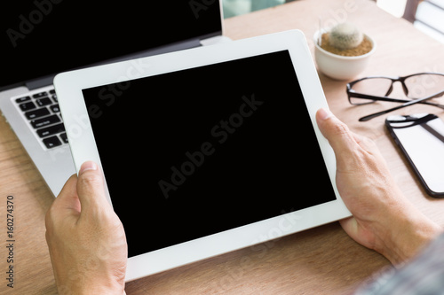 Hands holding dogital tablet pc black screen on wood table with laptop and smartphone for mockup advertising display design photo