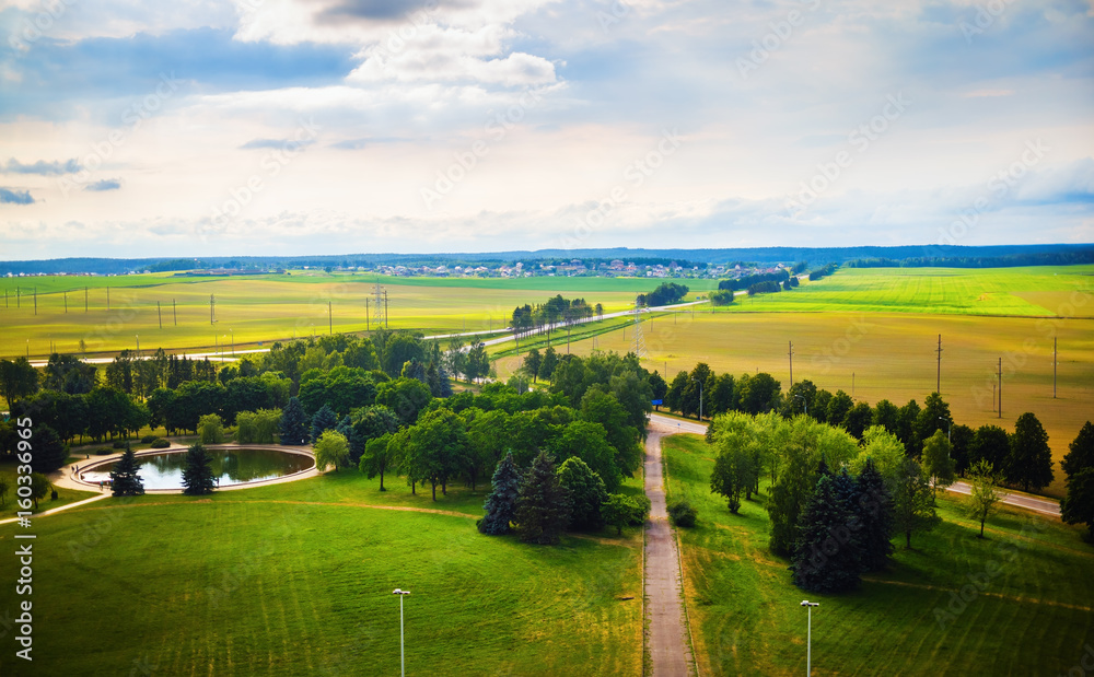 Obraz premium Green countryside landscape. Fields, meadows, trees, pond, roads, houses on the horizon and sky with clouds. Minsk region, Belarus
