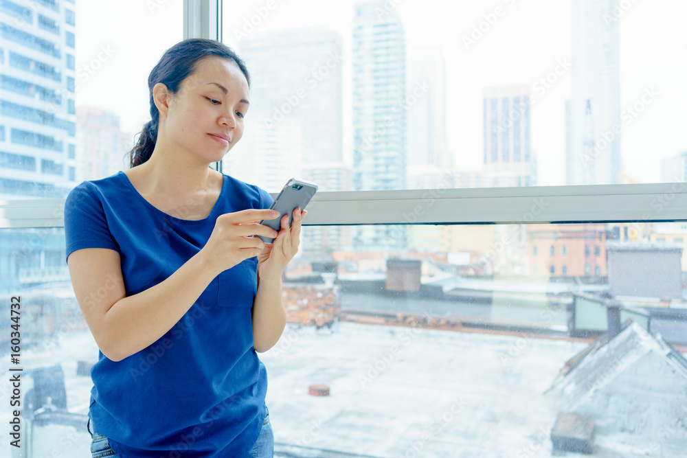 Fototapeta premium Woman standing indoors by window, day, urban scene, using mobile phone.