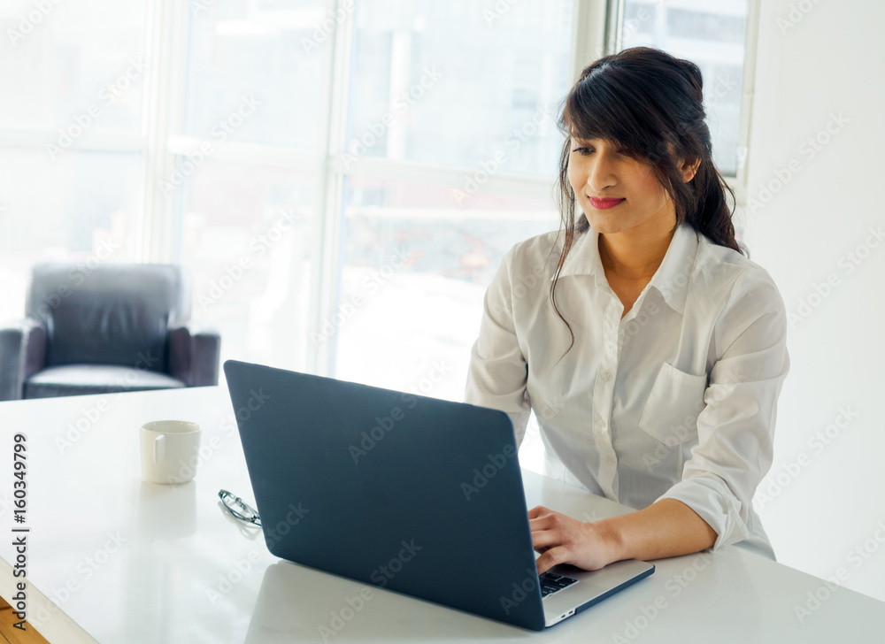 Young woman using laptop at desk, day, with window behind. Young woman using laptop at desk, day, with window behind.