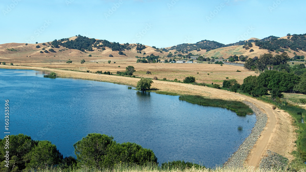 Panoramic view of the North side of the Lagoon Valley Park lake in Vacaville, California, USA, featuring the chaparral in the summer with golden grass and interstate-80 in the distance