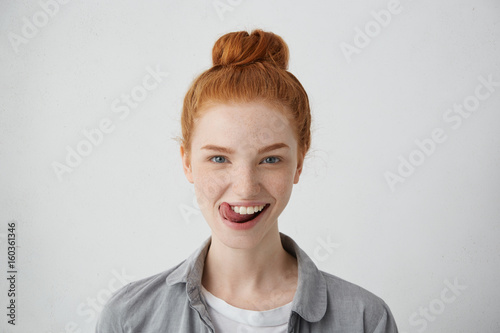 Young teenage female with ginger hair knot sticking out her tongue having funny look isolated over white background. Freckled student girl having fun being happy to finish her studying successfully photo