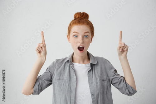 Astonished female with ginger hair bun, freckles and blue eyes pointing upwards with her fingers being surprised to see something. Concerned red-haired woman with eyes popped out and wide opened mouth photo