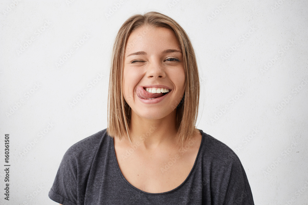 Horizontal portrait of optimistic funny girl with bob hairstyle showing her tongue and blinking eyes while posing against white studio background. Emotional positive young female making grimace Horizontal portrait of optimistic funny girl with bob hairstyle showing her tongue and blinking eyes while posing against white studio background. Emotional positive young female making grimace
