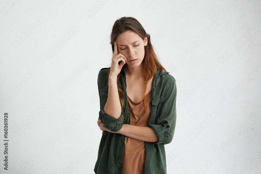 Exhausted and tired woman with dark hair standing against white wall with her eyes closed holding index finger on temple thinking about something. Upset female with tired expression. People and work