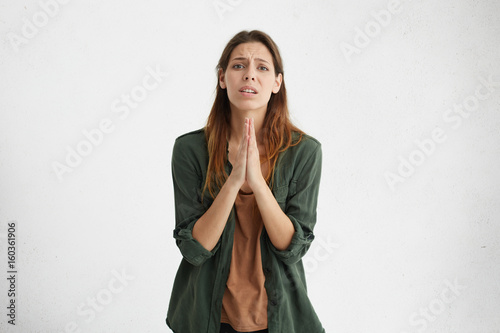 Worried apologetic brunette woman having some difficulties pressing her palms together begging for apologize. Apprehensive female expressing her anxiety asking God for health isolated over white wall photo