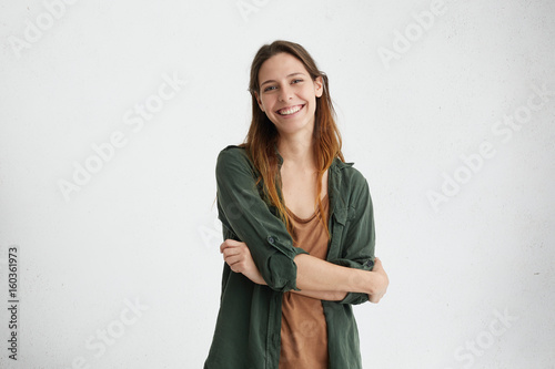 Photo of restful woman with dyed hair standing crossed hands having sincere and delightful smile demonstrating her perfect teeth posing over white studio wall. Happy female rejoicing her life photo