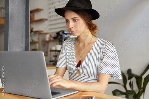 Young female freelancer with beautiful appearance wearing elegant black hat, shirt and pendants on neck sitting indoors over cozy interior working with her computer typing having serious look