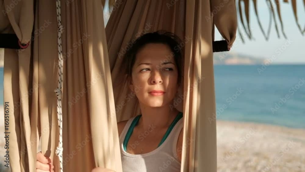 On the beach, a woman sits in a swinging hammock and looks into ...