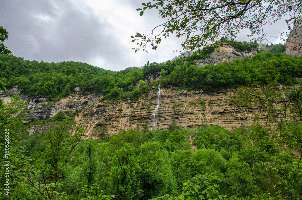 Waterfall on a high rock mountain. Cloudy landscape wallpaper with green Caucasus forest. Guam Gorge