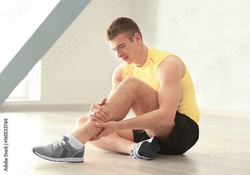 Young man suffering from pain in leg while sitting on floor indoors