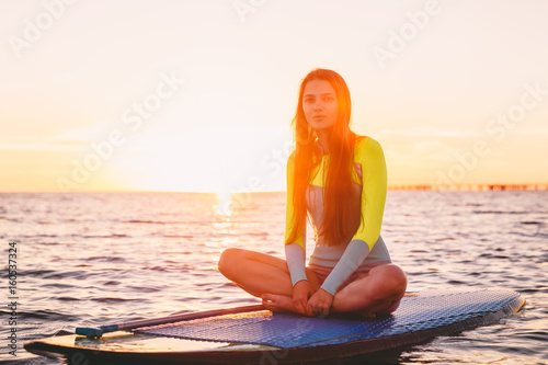 Young beautiful girl relaxing on stand up paddle board, on a quiet sea with warm sunset colors.