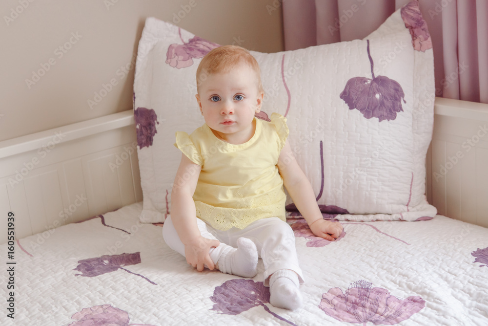 Portrait of cute adorable white blonde Caucasian smiling baby girl with large blue eyes in yellow shirt, sitting on bed, looking in camera Portrait of cute adorable white blonde Caucasian smiling baby girl with large blue eyes in yellow shirt, sitting on bed, looking in camera