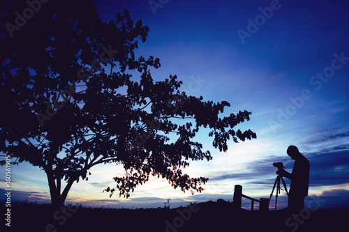 Silhouette photographer with camera on tripod taking pictures of nature tree on sunset.
