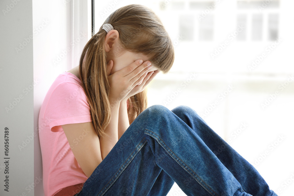 Sad little girl sitting on window sill at home Sad little girl sitting on window sill at home