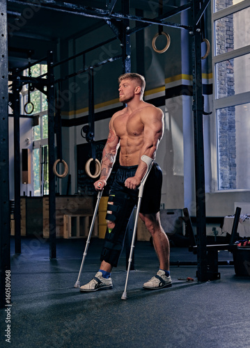 Shirtless bodybuilder on crutches near cross fit stand.