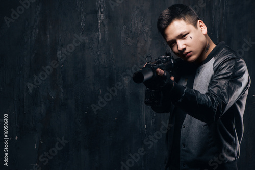 Sniper with tear tattoo on face and rifle close-up. Armed asian killer man with weapon and tattoo on dark background with free space. Outlaw, ghetto, murderer, contract murder, robbery concept © Photodrive