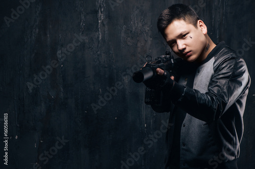 Sniper with tear tattoo on face and rifle close-up. Armed asian killer man with weapon and tattoo on dark background with free space. Outlaw, ghetto, murderer, contract murder, robbery concept