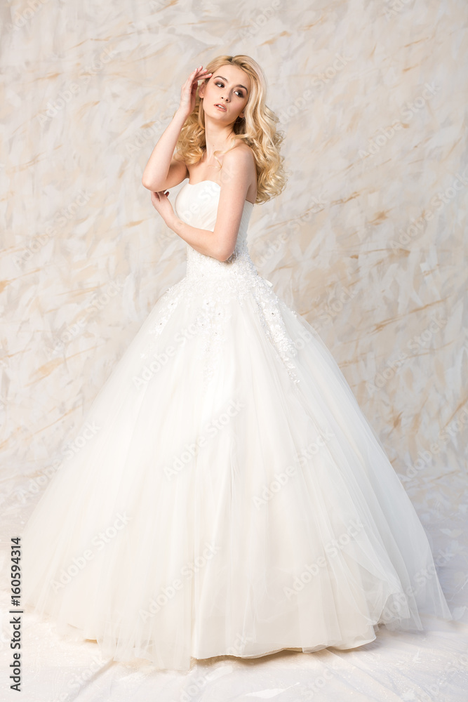 fashionable gown, beautiful blonde model, bride hairstyle and makeup concept - young lovely girl in white wedding festive dress, standing indoors on light background, romantic slender woman posing