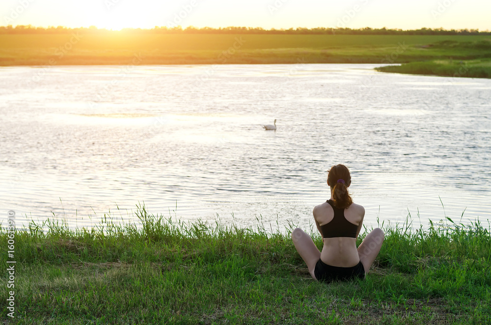 Young woman meditating in lotus position on the river bank, sunset. A woman with a healthy lifestyle, a copy of the free space.