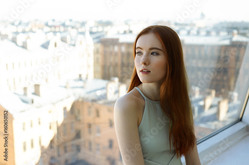 Portrait of beautiful redhead female student in cafe on the top floor and looks through the window at the city waiting her boyfriend. Attractive young woman looking away with dreamy expression