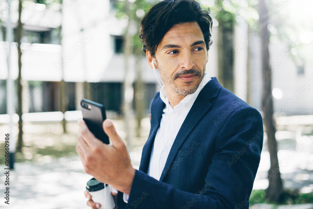 Closeup view of h andsome smiling businessman using smartphone for listining music while walking in city park.Horizontal,blurred background. Closeup view of h andsome smiling businessman using smartphone for listining music while walking in city park.Horizontal,blurred background.