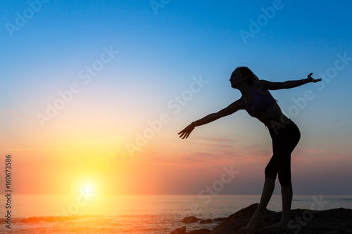 Silhouette of young flexible dancing woman on sea coast during twilight.