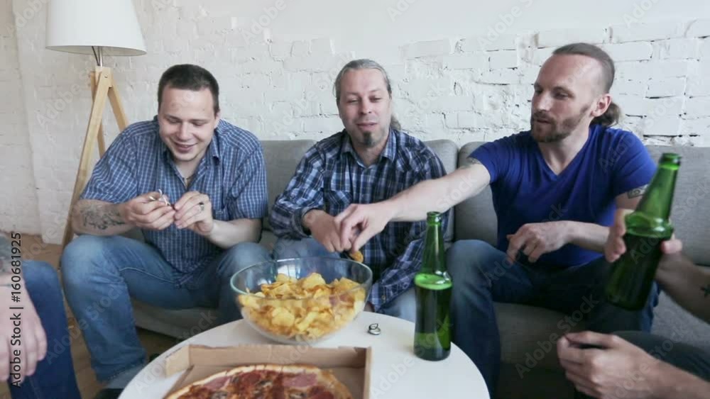 Group of men drinking beer, eating pizza, talking and smiling while resting at home on couch Group of men drinking beer, eating pizza, talking and smiling while resting at home on couch