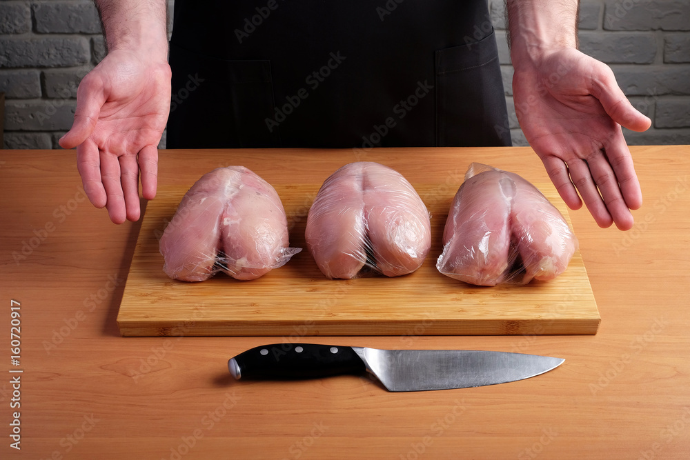 man in black apron holding open palms on the background wooden cutting boards and chicken fillet packed in plastic bags