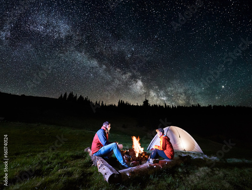 Night camping. Man and woman tourists sitting at a campfire near illuminated tent under beautiful night sky full of stars and milky way. Astrophotography