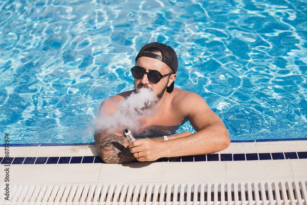 Men with beard in sunglasses and cap vaping in swimming pool. Stock Photo