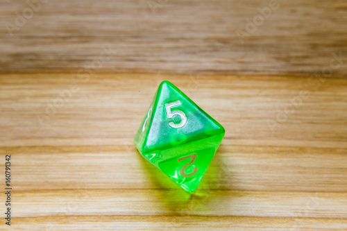 A translucent green eight sided playing dice on a wooden background with number five on a top