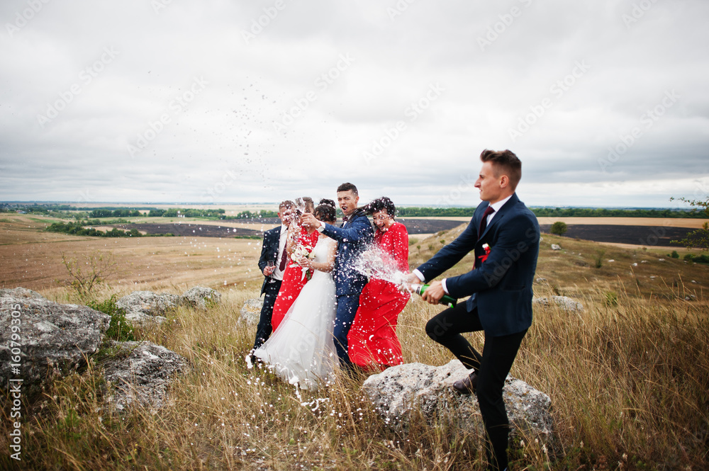 Groomsman or best man opening up the bottle of champagne in the countryside with wedding couple and braidsmaids with groomsman standing in the background.