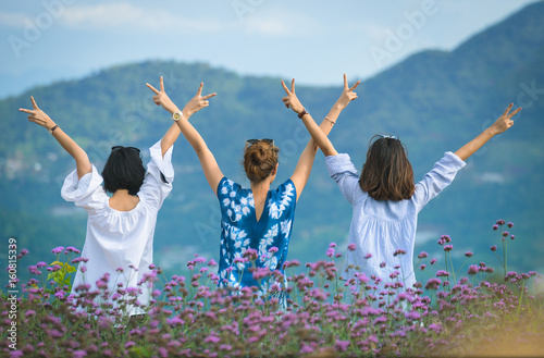 Rear view of Three teenage girls hands up staying in flower garden so happy with mountain background.