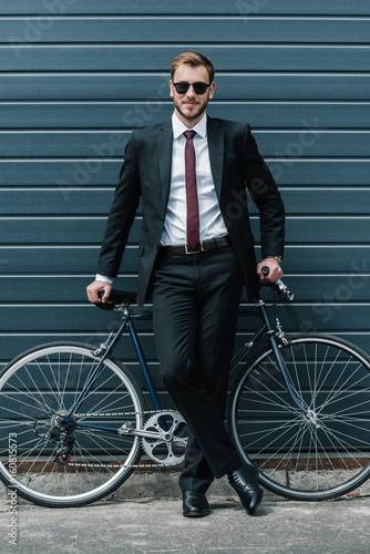 Stylish businessman in sunglasses sitting on bicycle and smiling at camera