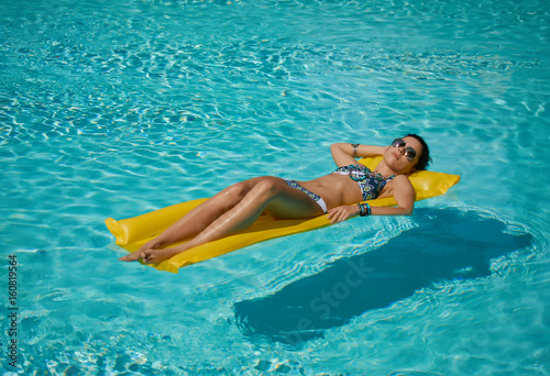 Young pretty fashion woman body posing in summer in pool with clear water lying on mattress in blue bikini and having fun
